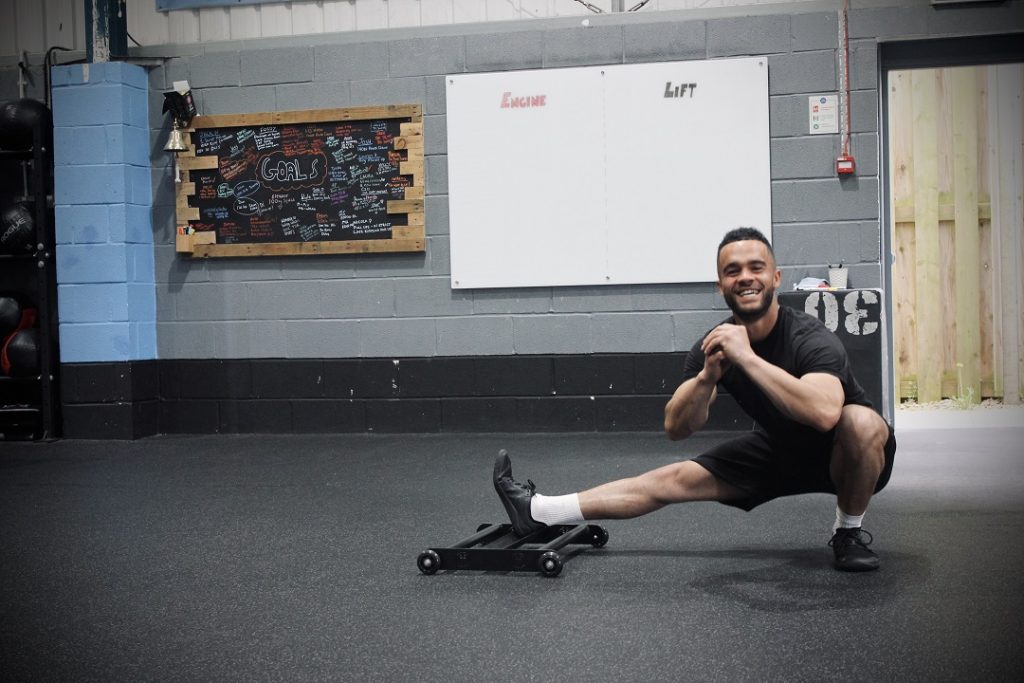 A smiling man using the Forte Fitness Roller. The man is in a lunged-squat position, with his right leg extended and his left knee bent, with his left foot flat on the floor.