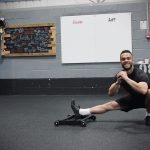 A smiling man using the Forte Fitness Roller. The man is in a lunged-squat position, with his right leg extended and his left knee bent, with his left foot flat on the floor.