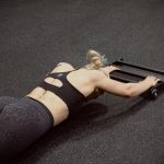 A female using the Forte Fitness roller in a gym setting. She is facing down in a plank position, with her arms extended using the roller.