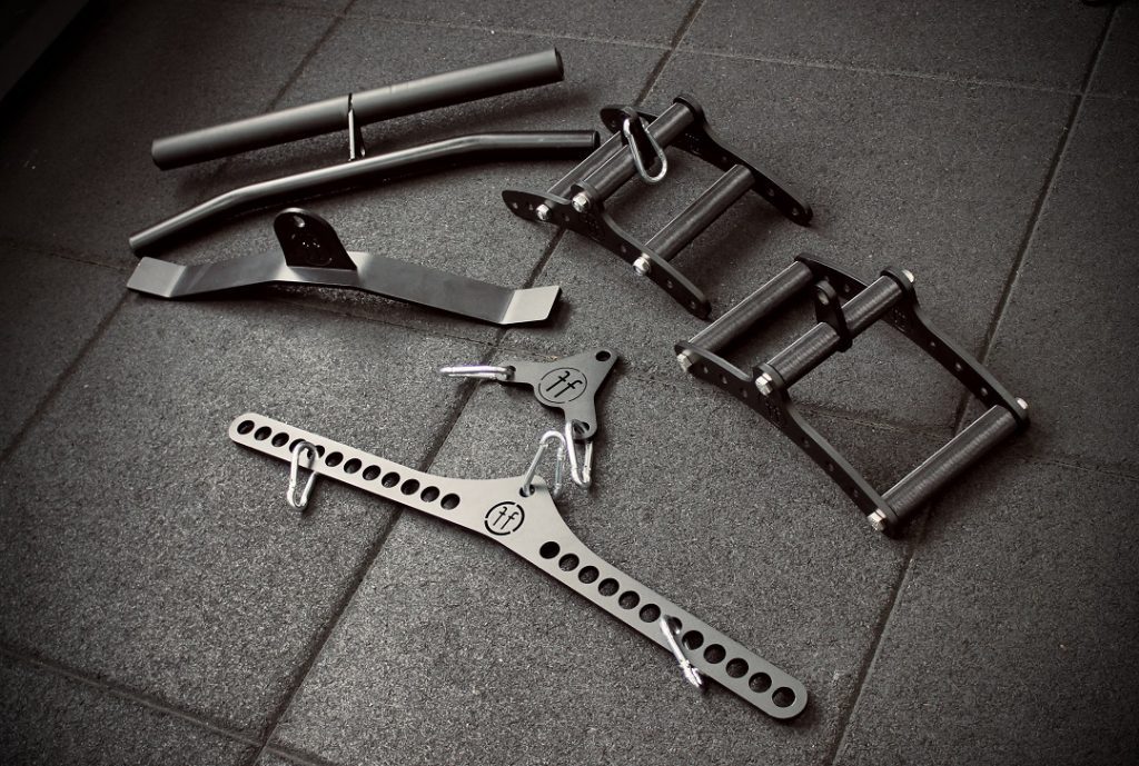 Forte Fitness cable attachments placed on the floor. From top left to bottom middle, this includes: a curl bar, 2 cable row attachments, a pushdown attachment, a pulldown attachment and an adaptable clip for weights.