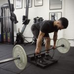 A man using the Forte Fitness step. He is stood on the lower step, and is bending over in a forward position while holding a bar with weights attached. His knees are bent to complete the deadlift maneuver.