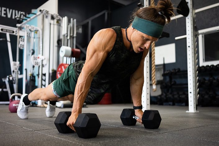 A male completing a plank on the floor. His hands are holding onto weights, which keep the front of his body lifted higher than the bottom half of his body.