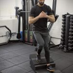 A man using the Forte Fitness step and incline boards. The incline board is placed on the top step, and the man is at the beginning of his squat, with his right leg placed on the incline board.