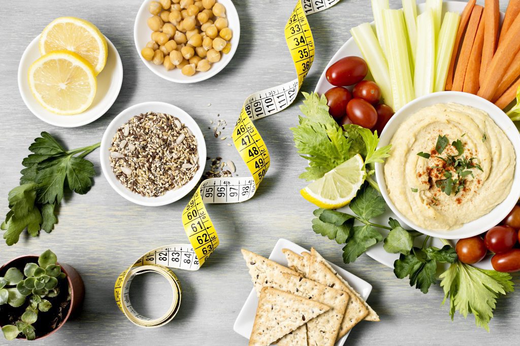 top view of a table with a bowl with 2 halves lemon, a bowl with chickpeas, a little indoor plant pot, some parsley, a tape measure, a plate with small slices of flat bread, a bigger plate with coriander, piccolino tomatoes, a bowl of hummus, celery and carrot sticks