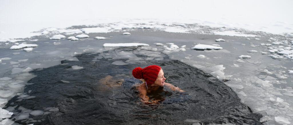 An active senior woman swimming in water hole in frozen lake outdoors in winter, cold therapy concept.