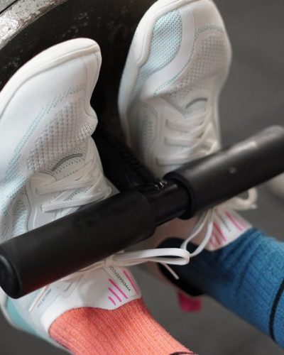 A Forte Fitness nordic bar attachment attached to a weight. A females feet are placed between the bar and the weight, demonstrating the foot positioning used for this movement.